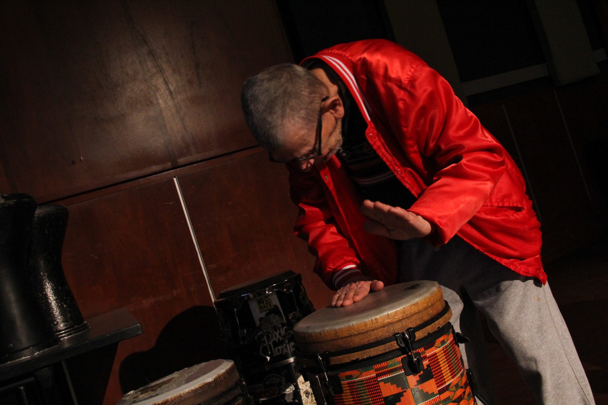 A man wearing a bright red jacket beats on a large drum.