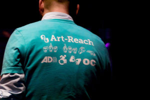 The back of Art-Reach staff member Charlie Miller, wearing an Art-Reach t-shirt with accessibility icons on it.