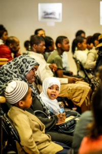 Two children sit with a woman as members of an audience.