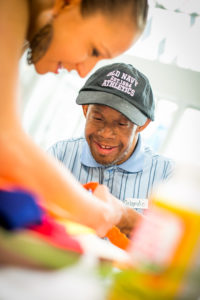 A man smiles as a woman helps him with his artwork.