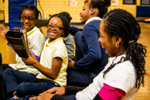 a group of girls laugh while one of them holds a large cowbell during a presentation by in-facility artist Tony Mascara.