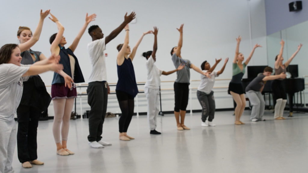 PA Ballet dancers and Saint Katherine Day School students stand in a gently curving line, with their arms outstretched in various positions.