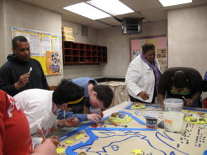 People surround and bend over a table with different small objects on it.