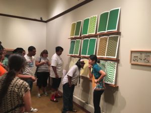 Art-Reach staff member Angela Wang gestures at adaptive tour attendees, while standing in front of shelves holding open books, printed with green patterns.