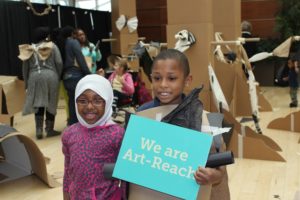 Two children, one girl and one boy, hold up a teal sign that reads "We are Art-Reach"
