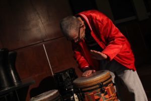 A man wearing a bright red jacket beats on a large drum.