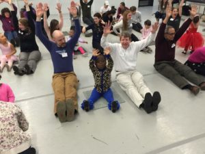 children and their parents hold their arms over their heads at a ballet lesson.