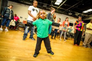 A child in a green shirt balances on his right foot, surrounded by smiling adults.