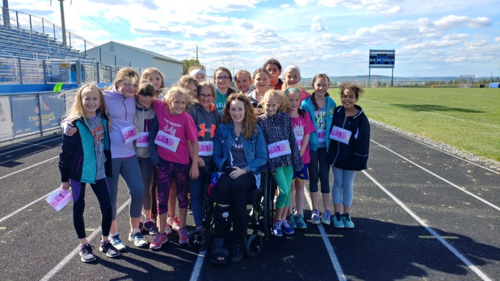 Girls on the Run Group pose in their running bibs on the track. In the middle of the girls, their teacher Sheila sits in a wheelchair and arm brace surrounded by their smiles