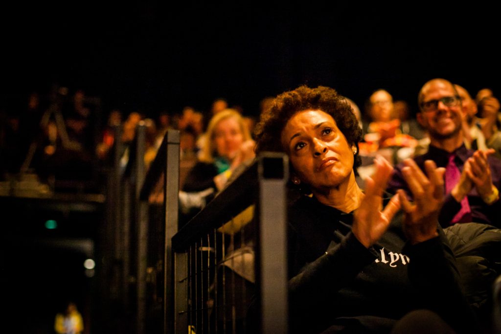 a view up the dark aisle of the theater while the audience looks on the stage and claps