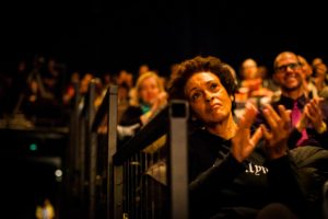 a view up the dark aisle of the theater while the audience looks on the stage and claps
