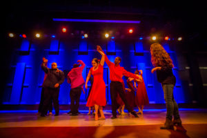 Mixed ability dancers performing on stage. In the center of the dancers is a woman in a red dress with her arm up getting twirled by a man in a matching red shirt