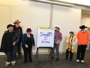 In Photo: Members in Halloween costumes pose with a large notepad that reads "StoryUP! with Art-Reach" Halloween costumes include, a person with a cape and top hat, sparkling jacket with red bow cap, sparkling zebra print jacket with a white safari hat, a hot dog, and a cowboy in a black and orange outfit.