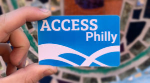 a hand holds a Blue and White card that reads ACCESS Philly with swirl logo.. behind the card is a blury mosaic wall