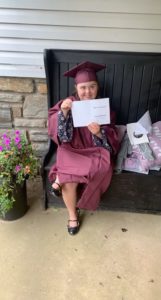 Muffy Tulskie, in a cap and gown, holds up a card celebrating her graduation day.