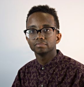 Headshot of Julian Harper. Julian self identifies as a Black male. In this image he has short hair, is wearing dark rimmed glasses and a dark patterned shirt. He is looking directly into the camera. 