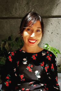 Reveca is a brown skinned woman with brown hair pulled back she smiles at the camera. wearing black shirt with roses print