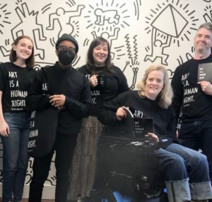 Art-Reach staff members wearing black shirts with white text that reads "Art is a human right" in front of a black and white Keith Haring mural featuring abstract drawings of people animals and objects.