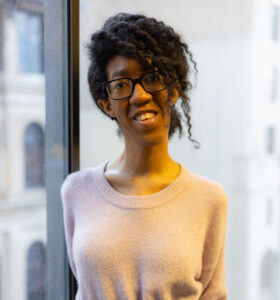 A black person with long coiled hair standing in front of a large window.
