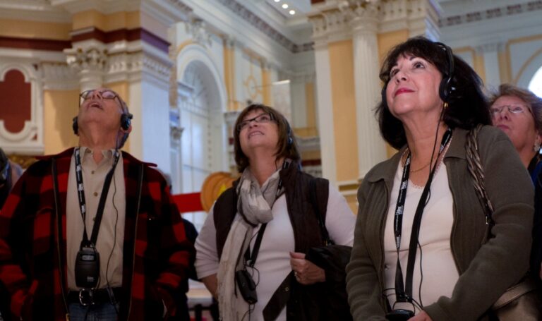 Three people experiencing an audiotour, wearing headphones and lanyards look upwards in a museum