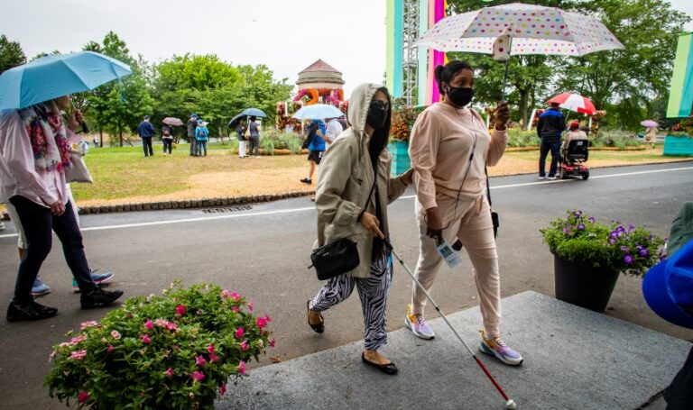 a person with an umbrella leading a blind person with a mobility cane across a small outdoor ramp in the rain