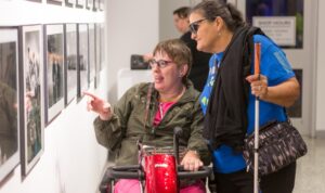 a mobility cane user and a person in a mobility scooter pointing to photographs at an art exhibit