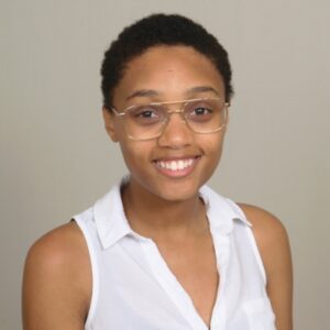 Alasia Destine-DeFreece, a person with brown skin, hair with short coils, wearing glasses and a white top