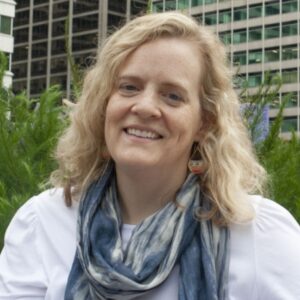 Katie Samson, a white woman in wheelchair shoulder length blonde hair with wearing a white shirt and blue and grey scarf