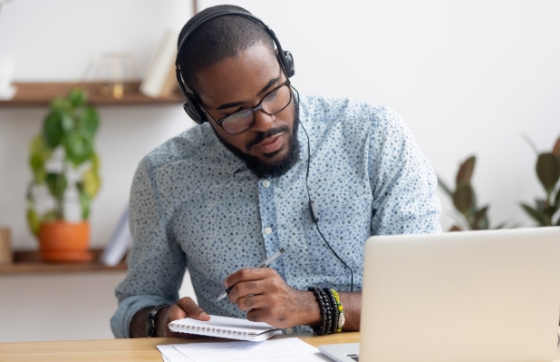 A black man with dark skin using a laptop and a notepad