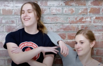 two young people with light skin, long brown hair standing in front of a brick wall in comfortable closeness