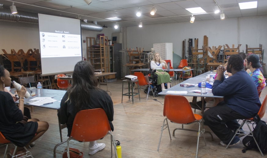 An art-reach staff member in a wheelchair provides training to a group of young adults in an art studio