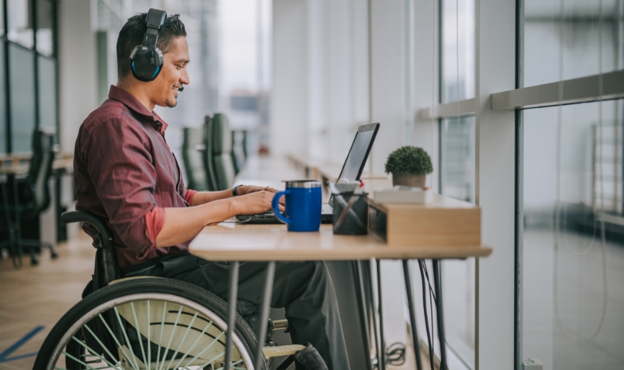 Asian indian male in wheelchair smiles while typing on a laptop and wearing headphone