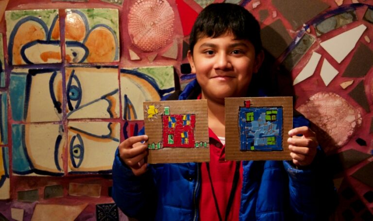 a child standing in front of a mosaic, holding two small artworks