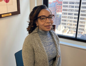 A woman smiling with brown skin and long black hair with caramel highlights wearing glasses, a knit blue turtleneck, and gray cardigan.