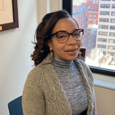 A smiling woman with brown skin and long black hair with caramel highlights wearing glasses, a blue knit turtleneck, and a gray cardigan.