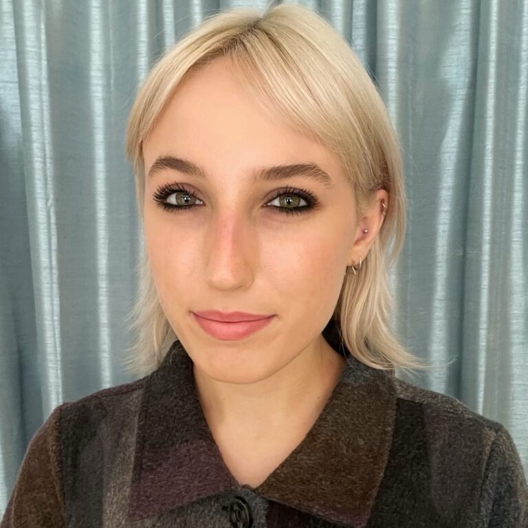 Avianna Miller, a young person with light skin, shoulder-length platinum blonde hair, and green eyes wearing a dark collared color-blocked shirt.