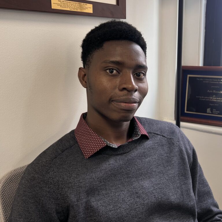 Munashe Mhlanga, a young person with black cropped hair, dark skin, and brown eyes wearing a black sweater over a maroon collared shirt.
